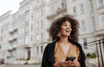 A person with curly hair, wearing large earrings and a brown top, stands outside an urban building, holding a phone and smiling.