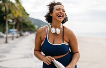 Plus size woman in fitness clothing stands on a beach promenade, with the ocean in the background. She is laughing happily, holding a water bottle, and enjoying a healthy workout lifestyle in summer.